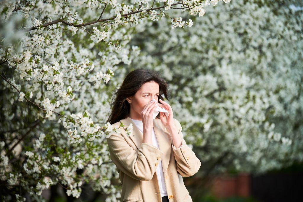 Mujer con alergia al polen limpiándose la nariz durante la primavera, rodeada de árboles en flor.