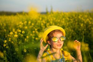 Niña con gafas de sol y sombrero amarillo en un campo soleado, protegida del sol.